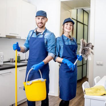 Portrait of a couple as a professional cleaners in uniform standing together with cleaning tools indoors